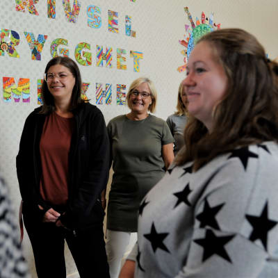 Några kvinnliga lärare står framför en skolvägg med texten trygghet, jämlikhet, trivsel. De ler och tittar på varandra. 