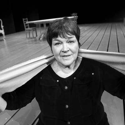 Teaterregissören Kaisa Korhonen står på ett stort trägolv på en teaterscen, runt halsen håller hon en scarf. 