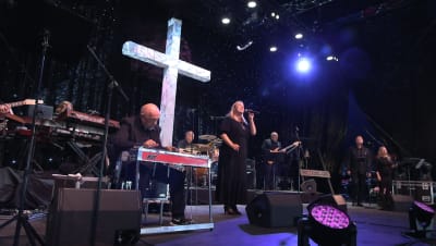 En kvinna som sjunger, en man som spelar keyboard, två körsångare och en elgitarrist står på en scen med ett stort vitt kors i bakgrunden.