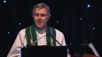 En präst klädd i vit kåpa med grönt band runt nacken. Prästen står vid en mikrofon på en scen.