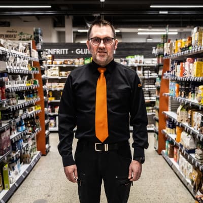 Man i mörk skjorta och orange slips står mellan butikshyllor