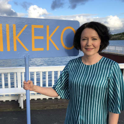 Rouva Jenni Haukio pitelee Unikeko-kylttiä, taustalla merimaisema.