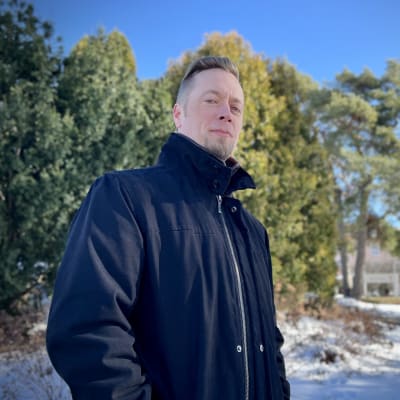 Turun reserviläiset ry:n Joose Olli musta takki yllään katsoo kohti kameraa, taustalla näkyy pensaita ja puita.