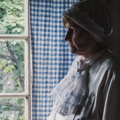 Kvinna i vit spetsklänning och vit klut på huvudet tittar ut genom fönster, som omgärdas av blåvitrutig gardin.