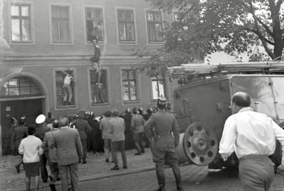 En svartvit bild från 1960-talet. Människor har samlats ute på gatan framför en byggnad och tittar på tre människor som försöker klättra in och ut genom fönster på väggen.