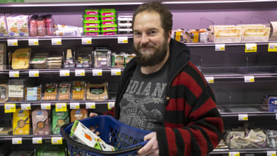 En man står framför kötthyllan i en mataffär. Han håller upp en butikskorg med varor i.