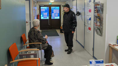 Sten Lindgren står inne i vårdcentralen. En äldre dam sitter på en orange stol.