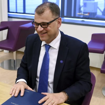 Juha Sipilä sitter bakom ett bord och skrattar.