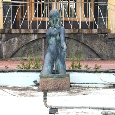 En staty, kvinnlig figur, i en liten ljusblå bassäng utan vatten, sand och löv och vattenrör syns på bassängbottnen.