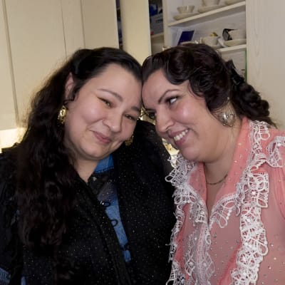 Kaksi romaniasuun pukeutunutta naista seisoo keittiössä ja hymyilee päät vastakkain.