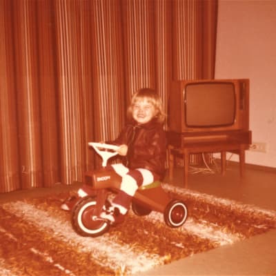Noin kolmevuotias Jenni Pääskysaari ajaa kotinsa olohuoneessa kolmipyöräisellä polkupyörällä 1970-luvun lopulla. 