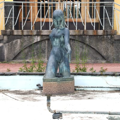 En staty, kvinnlig figur, i en liten ljusblå bassäng utan vatten, sand och löv och vattenrör syns på bassängbottnen.