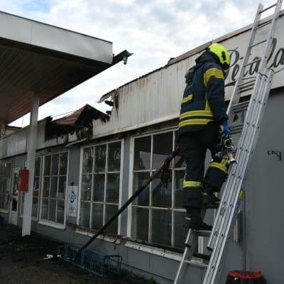 En bensinstation där taket brunnit. En brandman klättrar upp för en stege med vattenslang.
