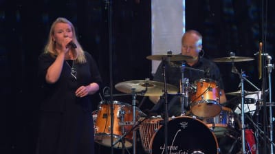 En kvinna i svart klänning sjunger i en mikrofon. En man i svarta kläder spelar trummor.