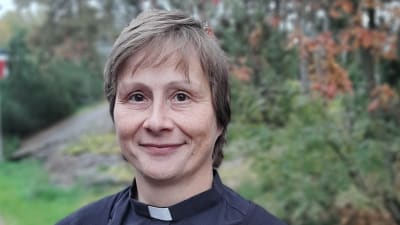 Saara Maria Roto, en dam med beige hår, svart klänning och prästkrage, tittar rakt in i kameran.