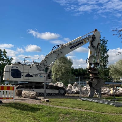 Työmaa-aita, betoninpaloja maassa, kaivinkone. Taustalla Kymijoki ja silta.