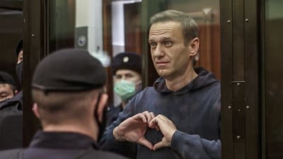 Aleksej Navalnyj står i en glasbur omgiven av poliser och formar ett hjärta med händerna.   