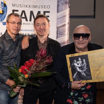 Remu Aaltonen, Ile kallio och Janne Louhivuori poserar med en blombukett och med en Hurriganes-plansch. Remu har solglasögon på sig. 