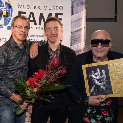 Remu Aaltonen, Ile kallio och Janne Louhivuori poserar med en blombukett och med en Hurriganes-plansch. Remu har solglasögon på sig. 