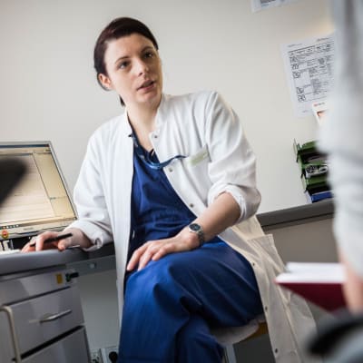 En läkare i uniform sitter på en stol vid en dator och pratar med en patient som syns i förgrunden.