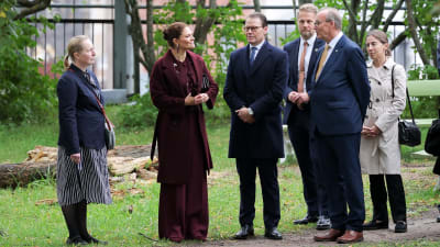 En grupp människor, bland dem kronprinsessan Victoria och prins Daniel, står på en stig som går över en gräsmatta. I bakgrunden står en rad träd.