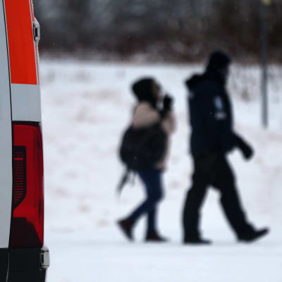 Rajavartiolaitoksen pakettiauton takakulma, jonka takaa häämöttää sumeana virkapukuinen poliisi saattamassa toppa-asuista laukkua kantavaa henkilöä lumisessa maisemassa.