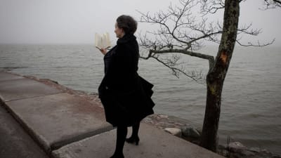 Kvinna står med bok i handen med havsutsikt i bakgrunden.