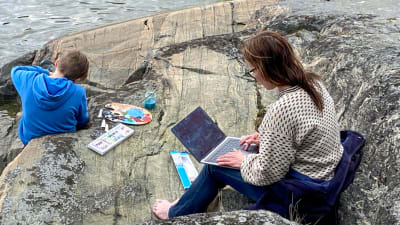 Kvinna sitter med bärbar dator på en klippa vid havet.