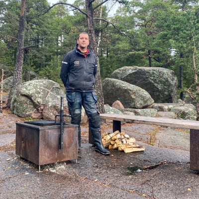 En man i mörka kläder står bredvid en grill och  några bänkar på en klippa.