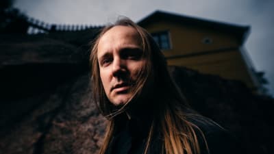 Kirjailija Mike Pohjola seisoo kallion edessä, takaa häämöttää puutalo.