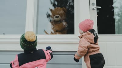Två små barn klädda i utekläder kikar in genom ett fönster där en teddybjörn sitter.