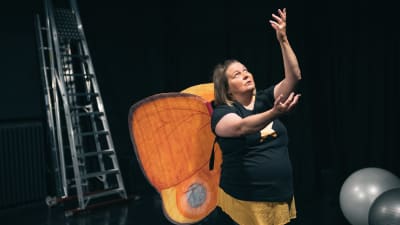 En kvinna med ett par väldiga orange fjärilsvingar på ryggen håller händerna teatraliskt uppsträckta.