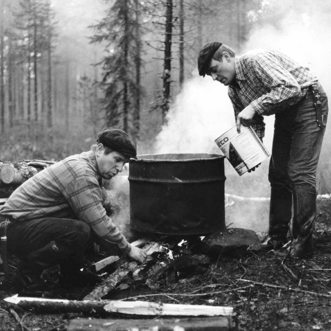 Kaksi lippalakkipäistä miestä puuhastelee metsässä öljytynnyristä tehdyn viinapannun äärellä: yksi kohentaa tulta, toinen on kaatamassa maalipöntöstä lisää nestettä pannuun.