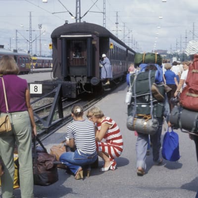 Helsingin päärautatieasemalla ihmisiä takaa päin matkatavaroiden kanssa. Kahdella rinkat selässä. Taustalla joku nousee junaan.