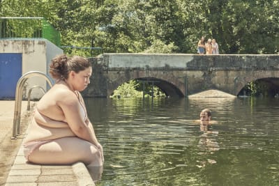 Sara (Laura Galán) sitter ensam vid en poolkant och ser ledsen ut, i bakgrunden syns tre flickor på en bro och i vattnet ser man huvudet på en man.