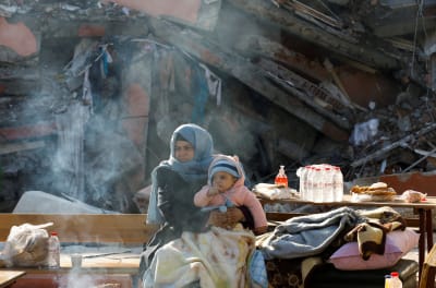 Ett litet barn i sin mammas famn, i bakgrunden syns ruiner efter jordbävning.