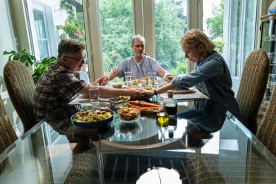 Tre vuxna personer sitter och håller varandra i händerna under en bordsbön vid ett middagsbord.