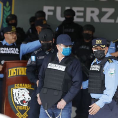 Juan Orlando Hernández i skottsäker väst, handbojor och munskydd, leds iväg av poliser.