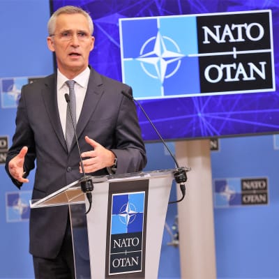 Stoltenberg puhuu. Hänen edessään on pieni puhujanpönttö, jonka päällä sojittaa kaksi mikrofonia. Stoltenbergillä on tummanharmaa puku ja vaaleanharmaa kravatti. Taustalla näkyy Naton logo ja tekstit NATO - OTAN.