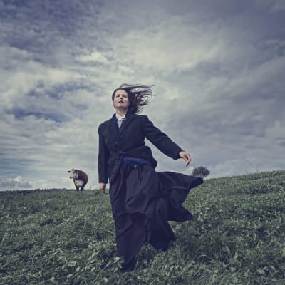 En kvinna i svart överrock står ute på ett blåsigt fält. I bakgrunden skymtar en ko. 