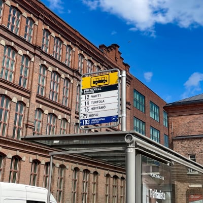 Frenckellin edustalla olevan bussipysäkin kyltit. Taustalla punatiilisiä rakennuksia ja sininen taivas.
