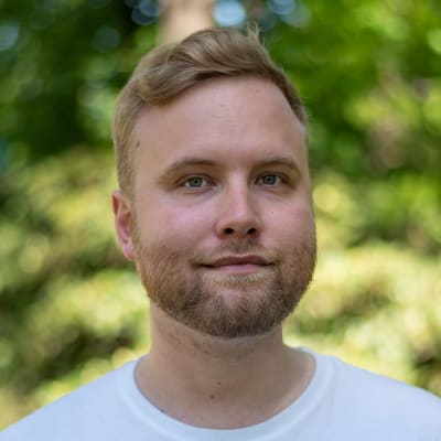 En man tittar in i kameran iklädd en vit t-skjorta, bakgrunden är ett ljusgrönt skogsparti.
