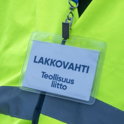 En person står med en reflexväst och en namnbricka runt halsen. På namnbrickan står det på finska att personen är en strejkvakt från Industrifacket.