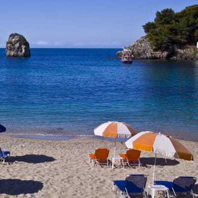 Bild på strand med blåvita och orangevita parasoll och solstolar.