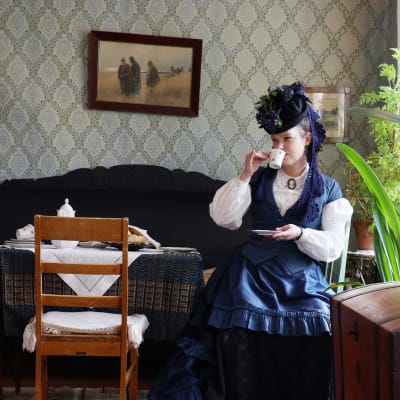 Taina Parikka juo teetä 1880-luvun henkinen leninki ja koristeellinen hattu yllään. Hän istuu Haminan Kauppiaantalomuseon salissa teepöydän äärellä. Huonekalut, tapetit ja taulut ovat 1800-luvun lopun tyyliä.