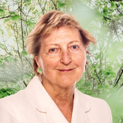 Ulla Tillander-Godenhielm småler in i kameran framför en grönskande bakgrund.