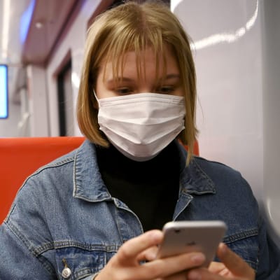 En ung kvinna sitter i metron i Helsingfors. Hon bär munskydd och tittar ner på sin mobil.