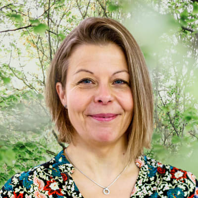 Anette Rönnlund-Nygård småler in i kameran framför en grönskande bakgrund.