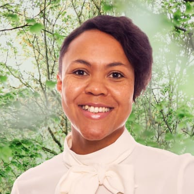 Elina Sagne-Ollikainen småler in i kameran framför en grönskande bakgrund.