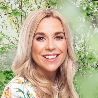 Krista Siegfrids småler in i kameran framför en grönskande bakgrund.